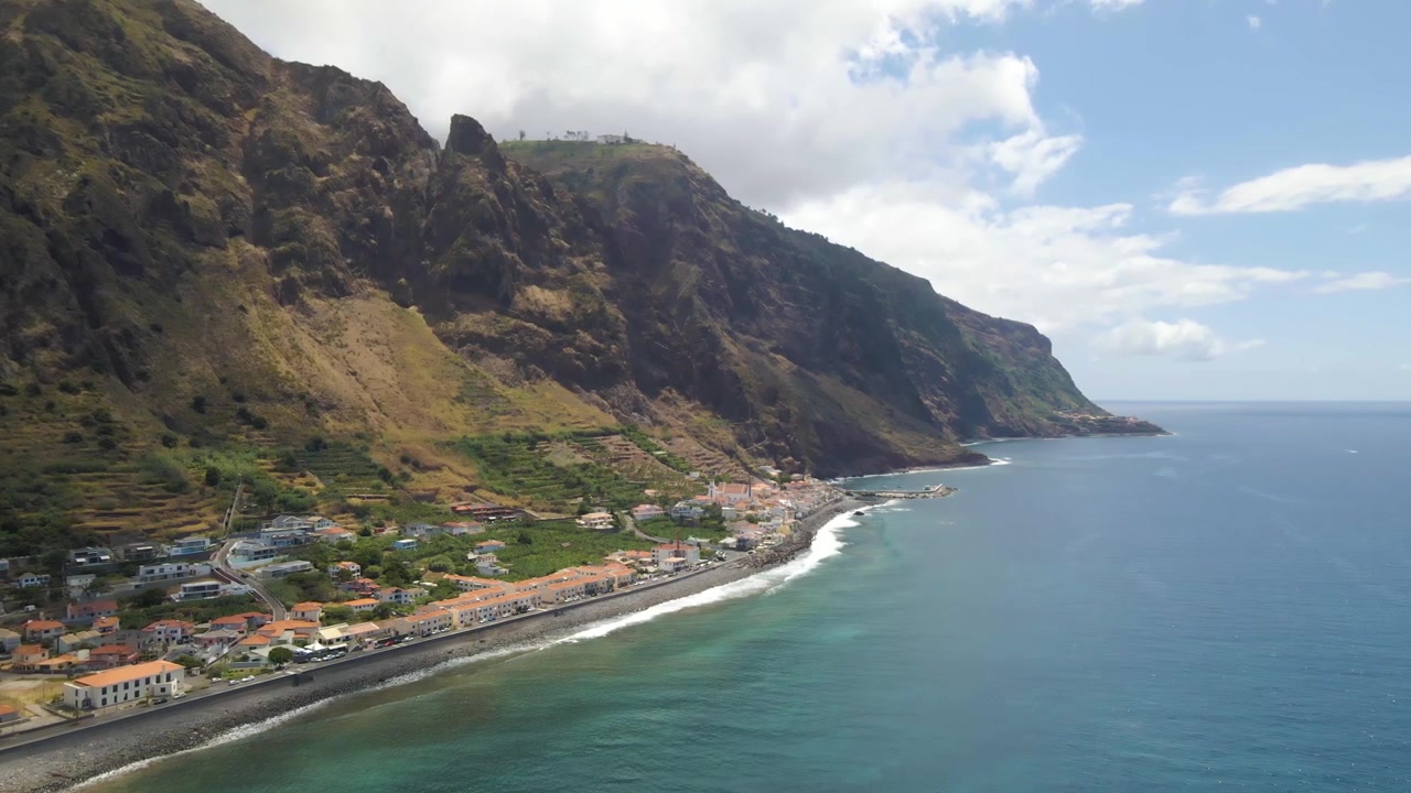 Riprese aeree che mostrano la disposizione completa del villaggio di Paul do Mar. L'insediamento compatto si estende lungo la costa sotto imponenti scogliere, illustrando la geografia unica dell'isola di Madeira