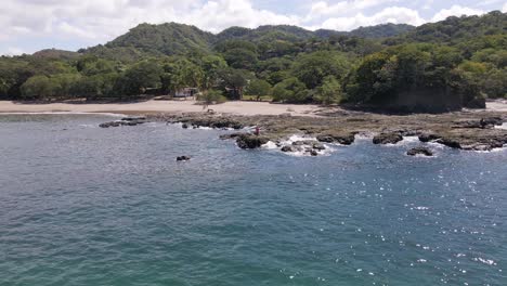 Toma-Aérea-Giratoria-De-Un-Joven-Parado-Justo-En-La-Costa-Siendo-Salpicado-Con-El-Agua-Del-Océano