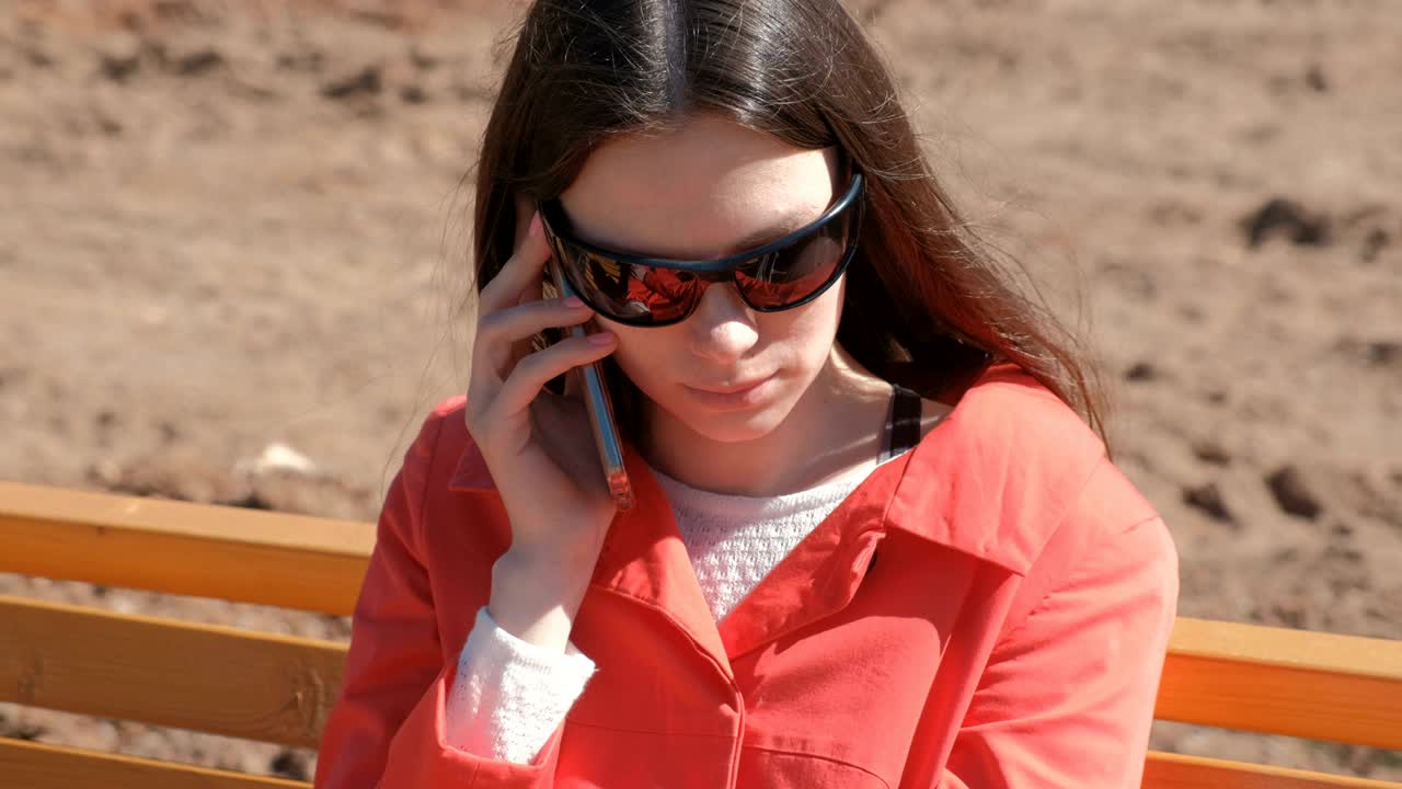 선글라스를 입은 젊은 갈색 소녀가 공원에서 벤치에 앉아 전화를 기다리고 있습니다.