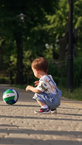 little boy squats near the ball and pushes it to roll. kid stands up and walks opposite side by the paved road. vertical video