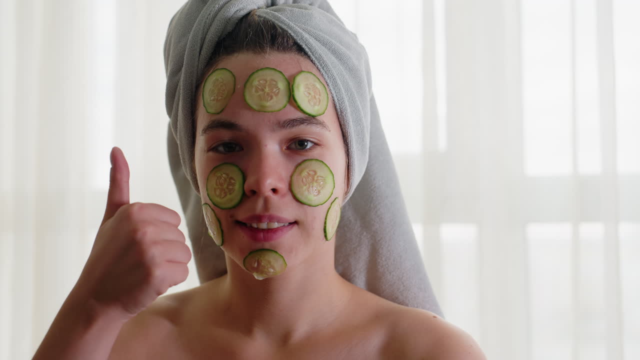 Portrait of woman with cucumber slices on face, hair wrapped in towel, giving thumbs up while enjoying relaxing skincare treatment, wellness, and rejuvenation moment with soft natural light