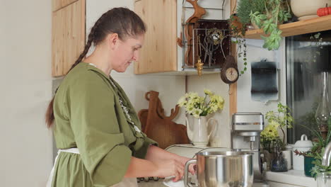 woman places glass cup on kitchen scale and carefully pours sugar inside for accurate measurement in bright, homey kitchen with decorative plants and utensils, wearing green dress and white apron