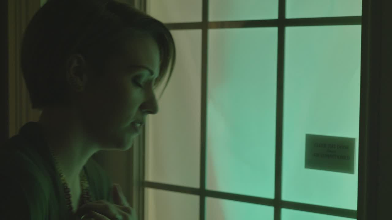 Scared woman is shaking and clutching a book. She hesitantly goes to open a glowing door with shadow figures behind it. She's super nervous and could be at a club