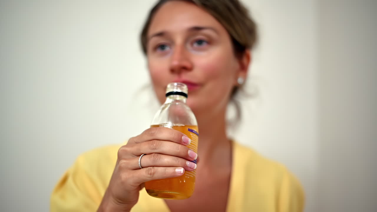 A woman takes a sip from a bottle, smiling and enjoying a refreshing drink. The setting is cozy and bright, perfect for a warm afternoon indoors