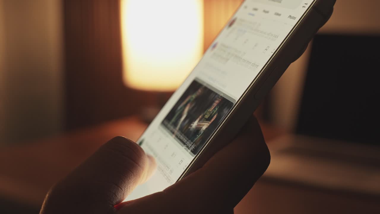 Hand holding smartphone and scrolling social media feed in dark room with desk on background, illuminated by warm tone light. Digital addiction and mindless content consuming