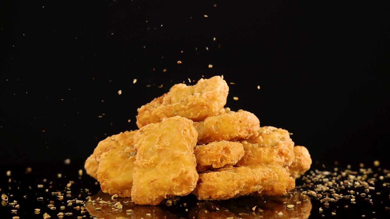 A pile of crispy chicken nuggets is sprinkled with seasoning against a black background, captured in slow motion with dramatic studio lighting and minimal camera movement