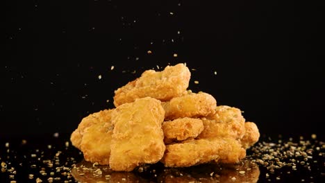 a pile of crispy chicken nuggets is sprinkled with seasoning against a black background, captured in slow motion with dramatic studio lighting and minimal camera movement