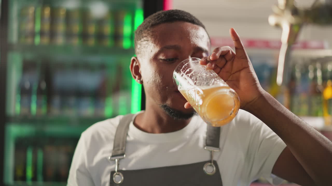 el barman bebe cerveza fresca en un pub de primer plano. el afroamericano toma un sorbo de una bebida alcohólica ligera y asente con la cabeza disfrutando de una calidad perfecta en el bar. el barman en el descanso
