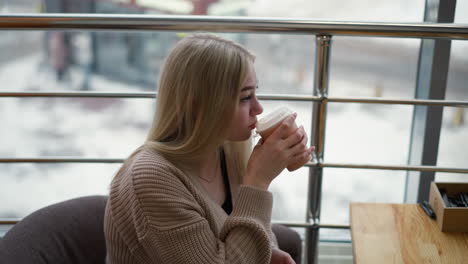 woman enjoys warm tea while gazing outside at serene winter cityscape, sitting in cozy indoor space with wooden table and modern decor, relaxed atmosphere with a peaceful urban background