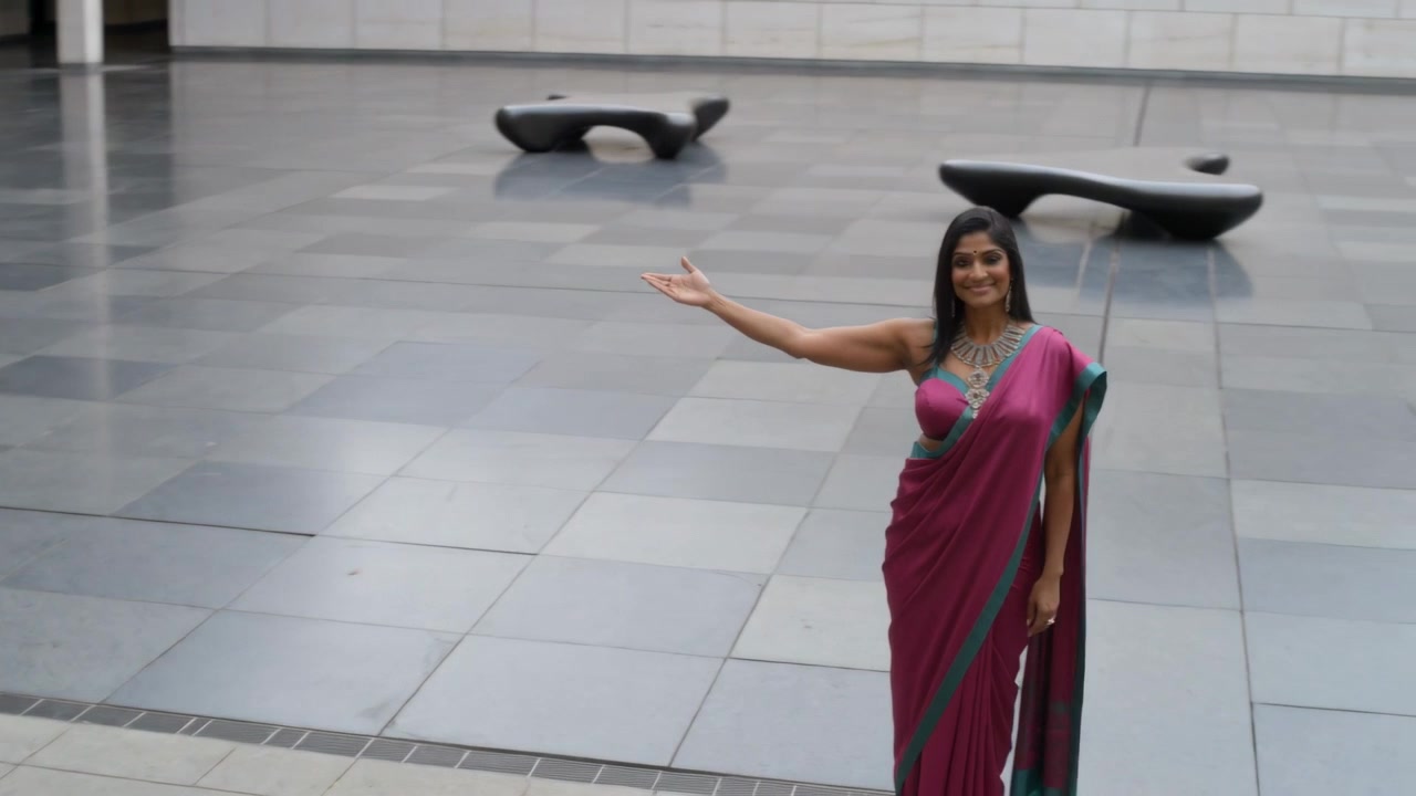 Una donna sorridente in sari magenta estende il braccio destro in una piazza cittadina, presentando uno spazio aperto, con spazio per il testo.