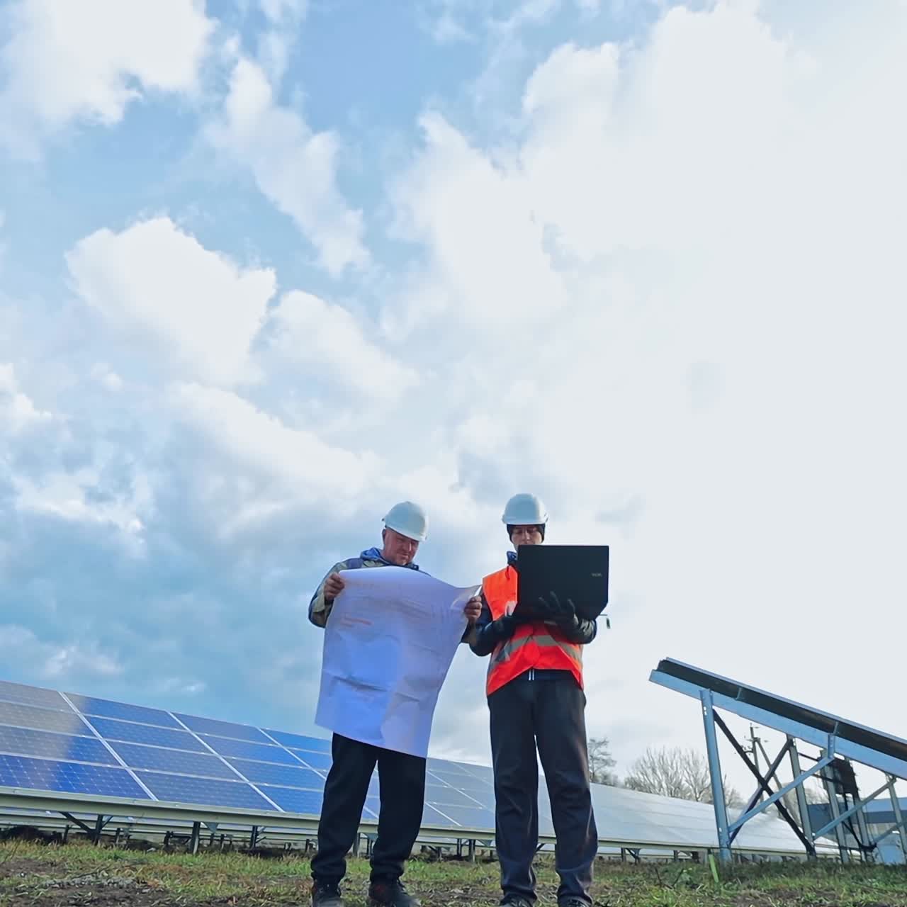 Engineers discussing blueprint on a laptop. Successful workers in protective uniform looking at plan on the background of solar panels farm.