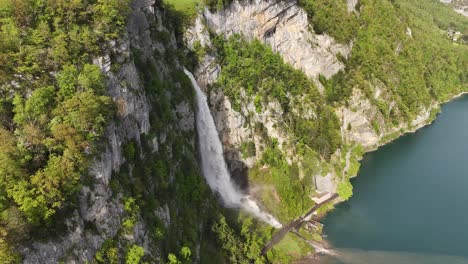 Aerial-view-of-Seerenbach-Falls-cascading-down-a-steep,-lush-cliff-into-the-blue-waters-of-Walensee,-surrounded-by-dense-forest-and-rocky-terrain-in-Amden,-Betlis,-Switzerland