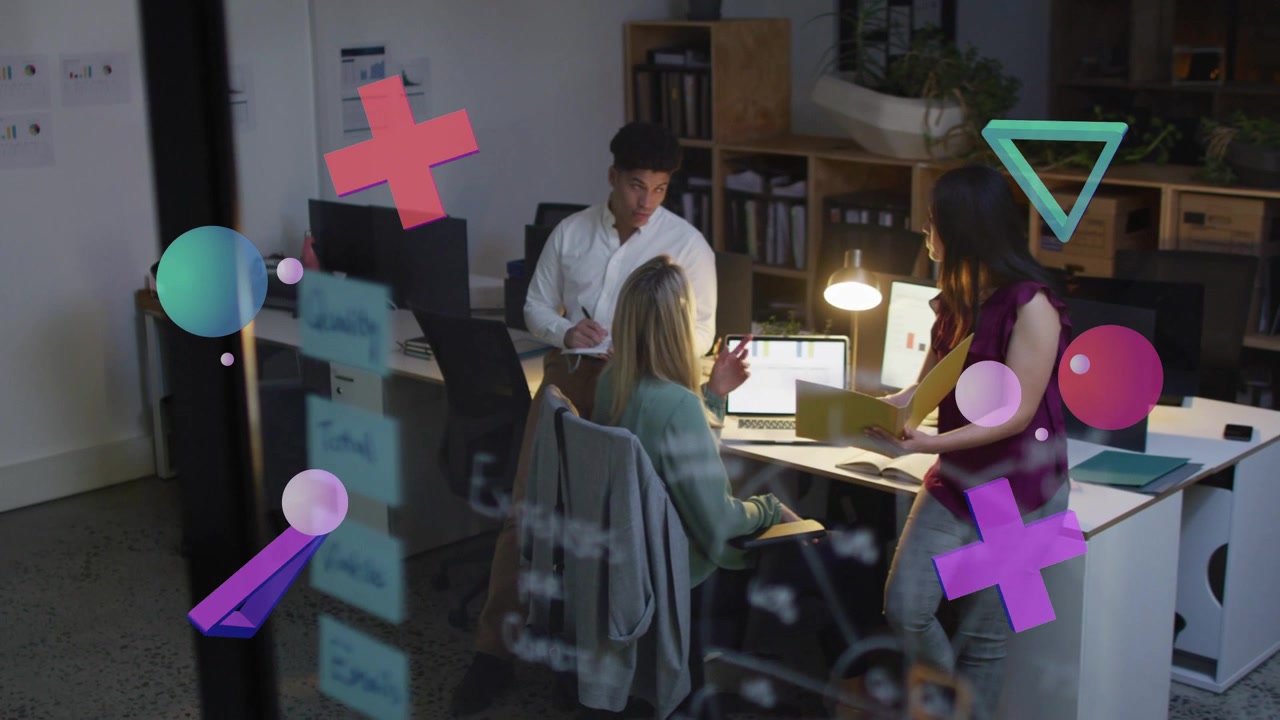 Kvinde ved bærbar computer peger, mens en kollega nærmer sig med en mappe, designteamet gennemgår layouts
