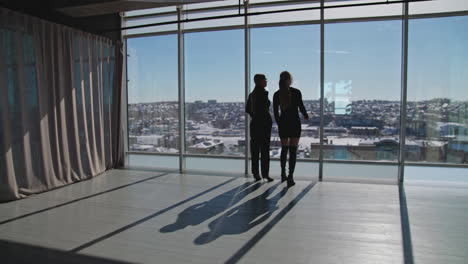 two girls wearing black clothes standing near the big windows in the spacious room. beautiful sunny cityscape at the background.