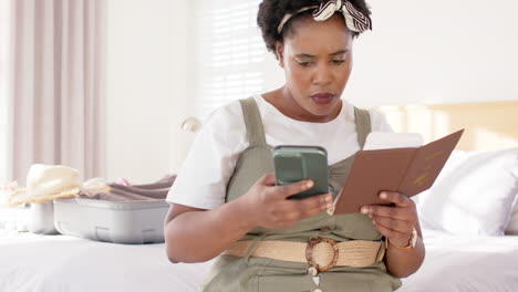 African-American-woman-looks-at-phone-with-concern,-passport-in-hand,-at-home,-booking-vacation