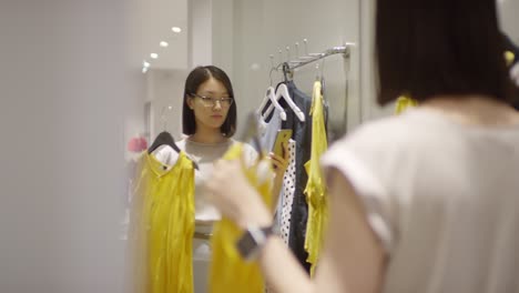 Smiling-Girl-Choosing-Clothes-In-Front-Of-The-Mirror-And-Taking-Photo-In-A-Shop