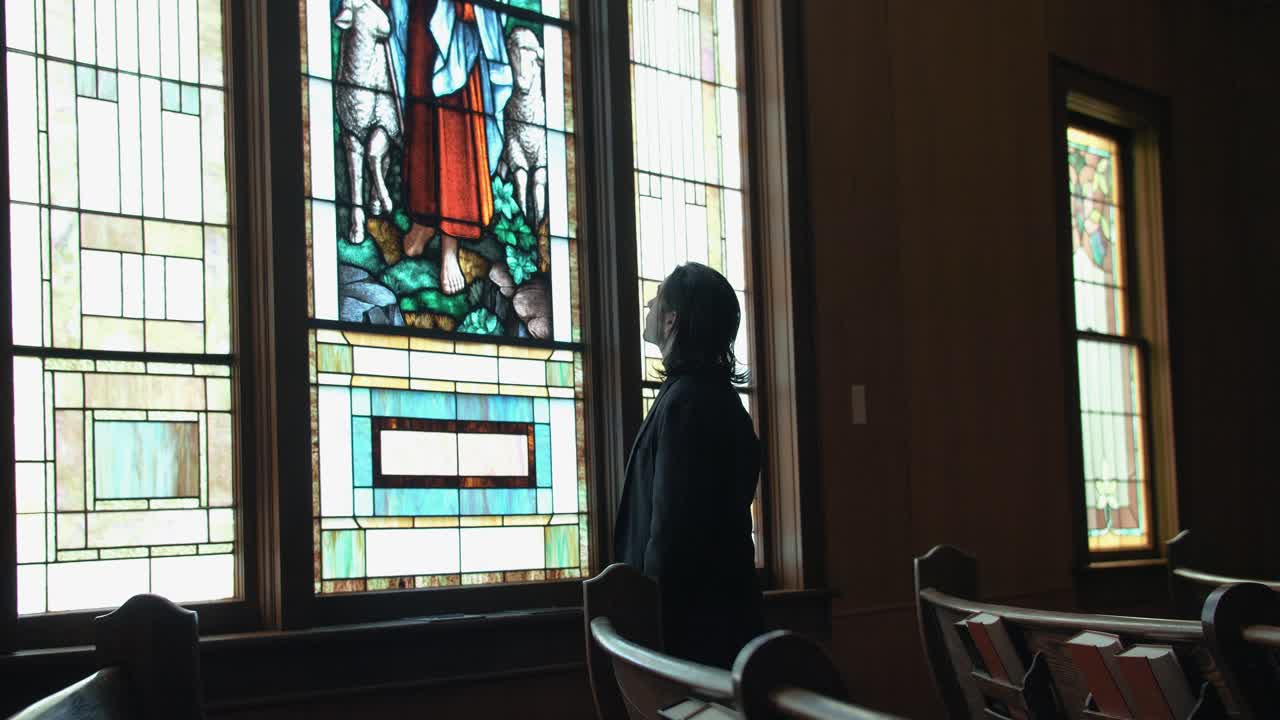 A man stands inside a church, gazing at a stained-glass window of Jesus the Good Shepherd, evoking themes of spirituality, faith, and quiet reflection