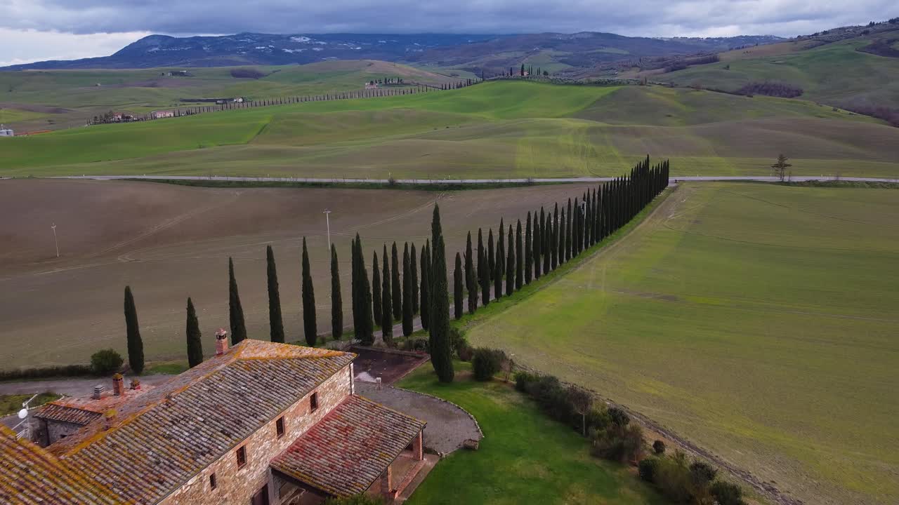 Capture a stunning aerial journey over Cinigiano, Tuscany, Italy. The drone glides forward, showcasing a charming farmhouse, cypress-lined roads, and lush hills under a dramatic sky.