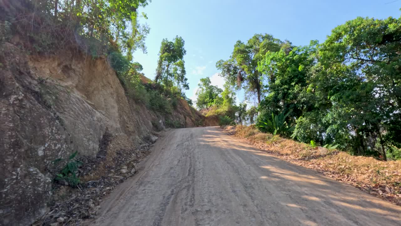 Vehicle travels uphill on winding dirt road, surrounded by lush tropical vegetation, bright daylight