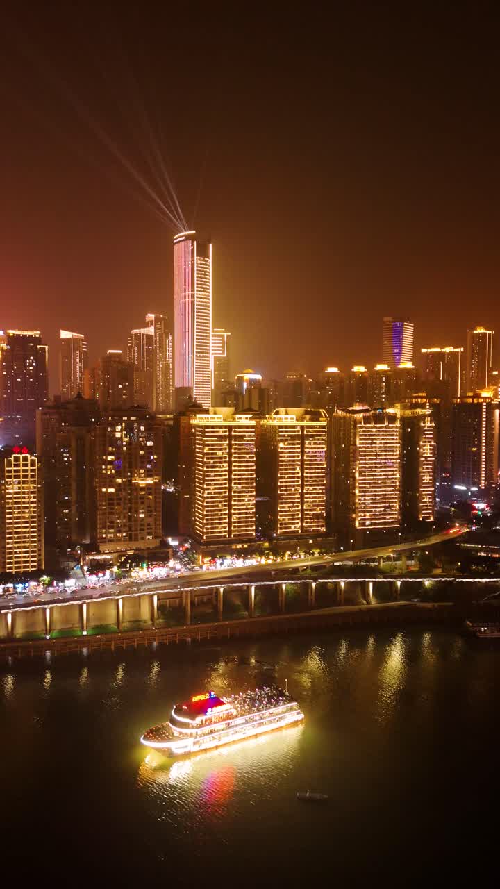 A vertical nighttime aerial view of Chongqing's illuminated skyline, featuring modern skyscrapers with colorful LED displays reflecting on the river below. China