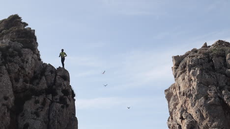 Un-Hombre-Comprueba-La-Altura-De-Un-Acantilado-Antes-De-Saltar,-En-El-Fondo-Gaviotas-Volando,-Cala-Llonga,-Ibiza,-España