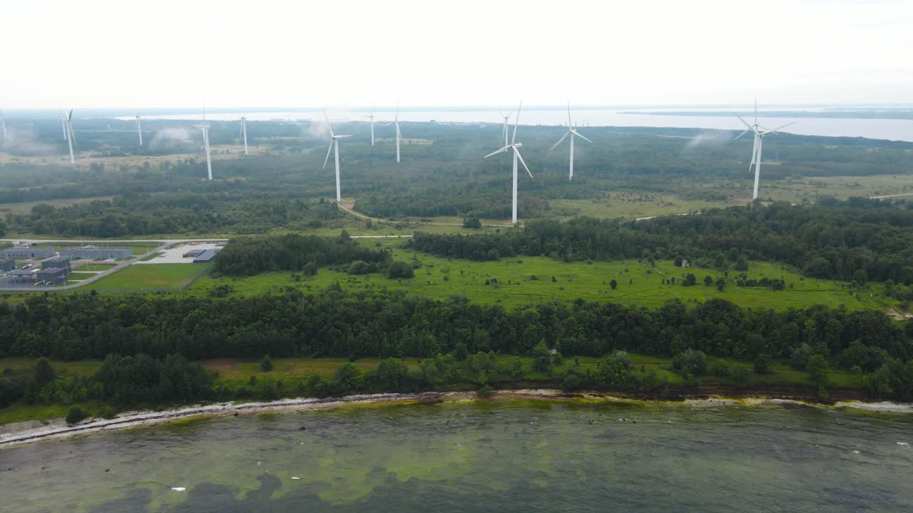 Aerial drone footage orbiting around a peninsula that has a wind farm on it with large and tall renewable energy creating wind turbines during a cloudy and misty day in Paldiski nature. Shoreline seen