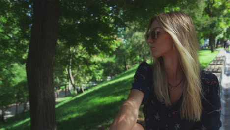 Young-Dreamy-Girl-Student-Wearing-Sunglasses-Lost-in-thought-Looks-Around-in-Shade-of-Trees,-handheld-shot