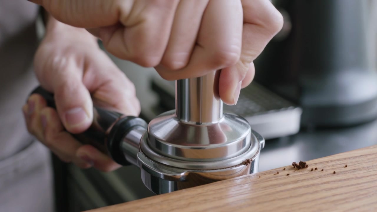 Mãos com mangas cinzas compactando, pressionando e girando o compactador em um porta-filtro dosado em um balcão de madeira para um expresso.