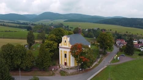 Vista-Aérea-De-Una-Iglesia-Rural-Amarilla-Con-Cementerio-Y-Colinas-En-El-Fondo-En-Bohemia