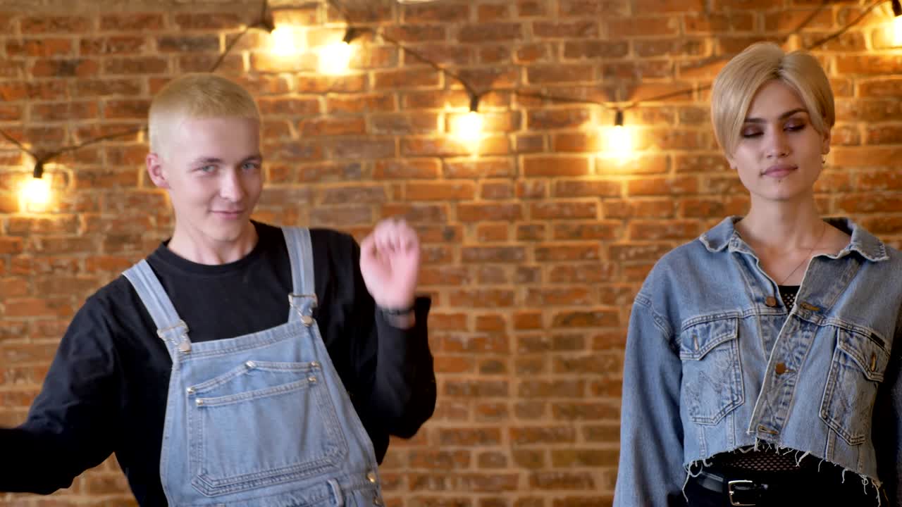 Young beautiful calmgirl is stending and extraordinary boy hipster is dancing next to her, brick background