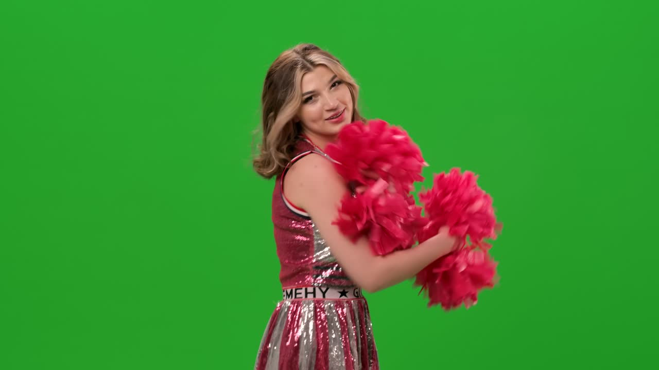 una adorable animadora en uniforme con pom-poms rojos realiza un baile jubiloso. retrato de una joven mujer sonriente bailando en el estudio en una pantalla verde. primer plano. cámara lenta