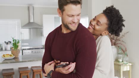 video of happy diverse couple hugging and using smartphone in kitchen