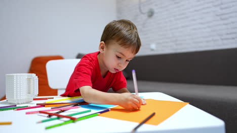 little toddler kid in red t-shirt sits at desk. colorful pencils are scattered in front of the child. baby boy picks a pen and draws on the paper.