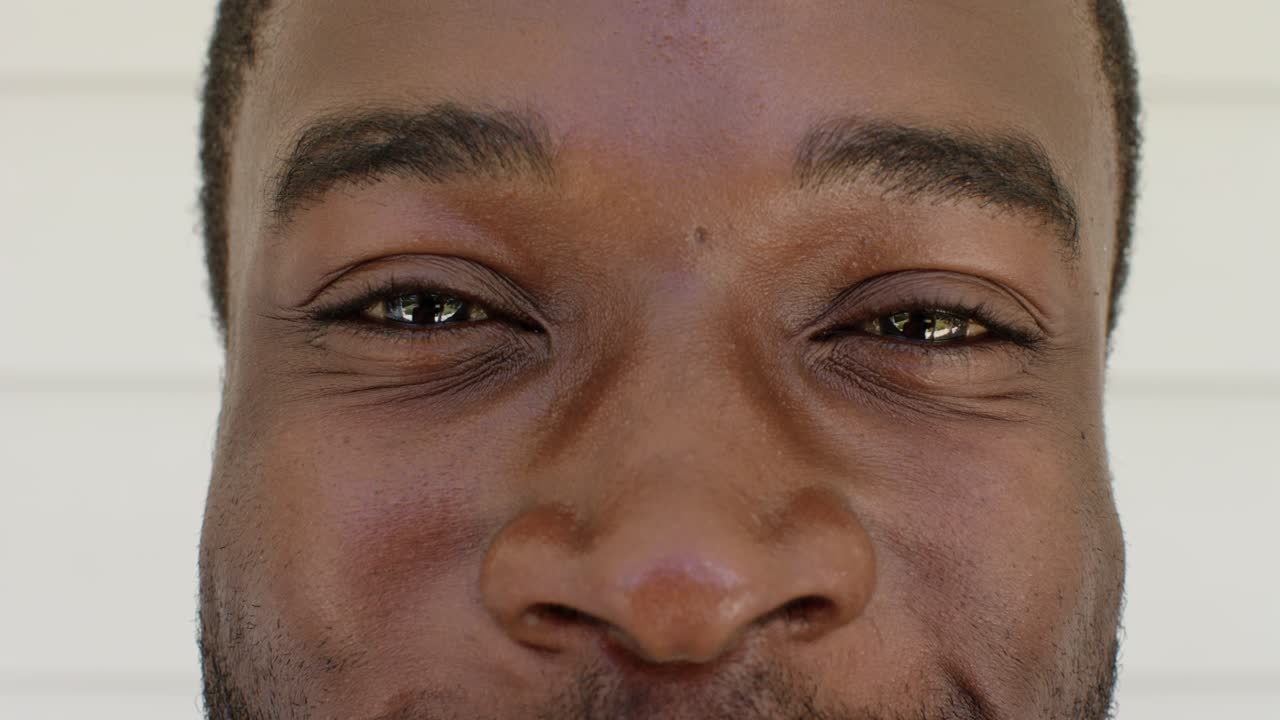 Adult African American male smiling and squinting while reacting to camera lens near pale siding
