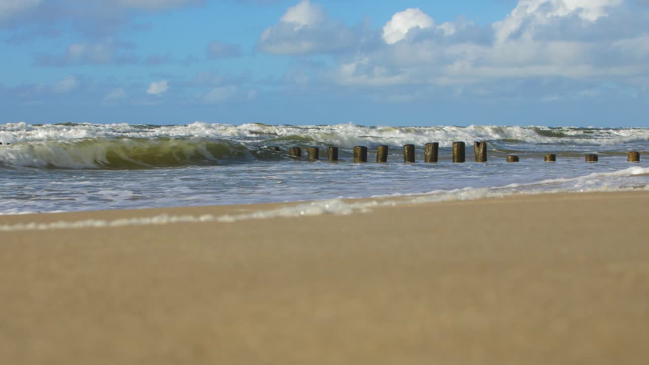 Stormy waves breaking against old wooden pier on the beach, white sand coast, sunny day, Baltic sea, distant low angle wide shot
