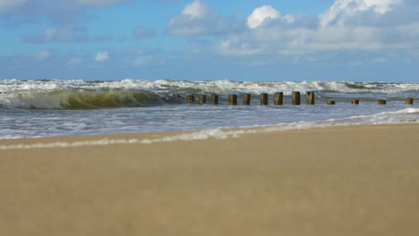 Stormy-waves-breaking-against-old-wooden-pier-on-the-beach,-white-sand-coast,-sunny-day,-Baltic-sea,-distant-low-angle-wide-shot