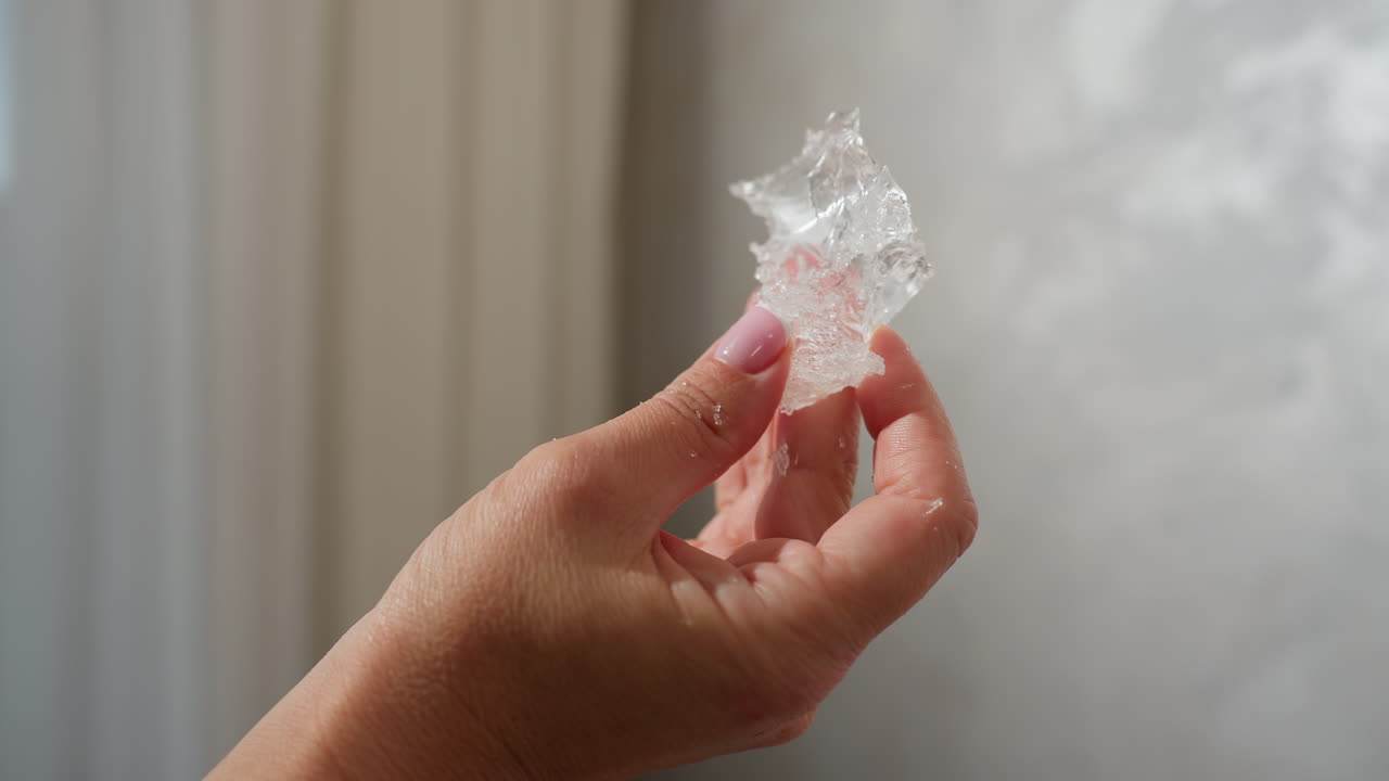 Close up of person carefully observing clear textured ice-like material held between fingers with soft pink nail polish against blurred neutral wall and soft natural lighting