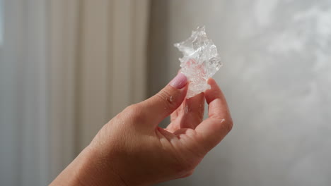 close up of person carefully observing clear textured ice-like material held between fingers with soft pink nail polish against blurred neutral wall and soft natural lighting