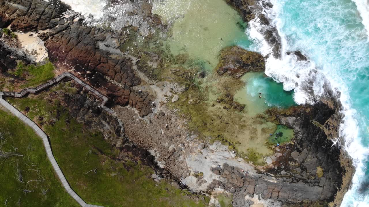 A drone swirls upwards capturing a stunning upward and spinning view of coastal rock pools, lush greenery, and crashing waves along the rugged shoreline at K'gari Island Champagne Rock Pools