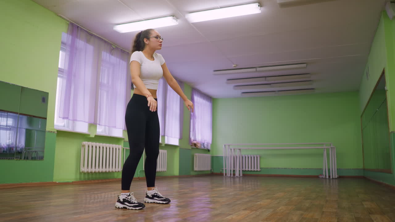 Lady standing in middle of hall painted green dancing with energy, wearing casual sportswear, sneakers, and glasses, moving gracefully under ceiling lights with wooden floor and large mirrors