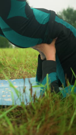 mujer de cabello gris acostada en una alfombra de yoga practicando la postura del puente con las manos apoyando su cintura, sentada en un vasto campo de hierba bajo un cielo nublado, con árboles en la distancia