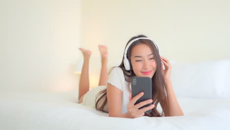 young thai smiling woman lying on bed with headphones listening to music while holding mobile phone