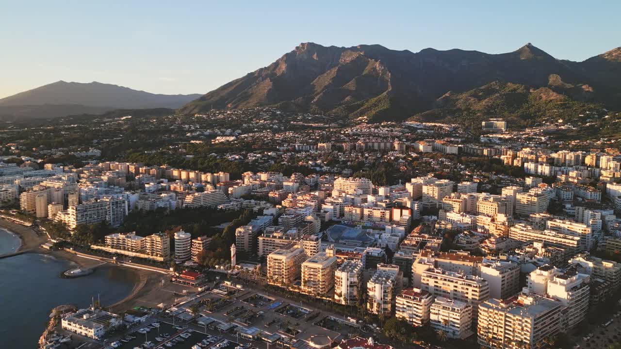 Sorvolo aereo con drone ad angolo elevato su Marbella, Spagna, e sul porto turistico al tramonto, skyline di lusso, con le montagne della Sierra Blanca sullo sfondo