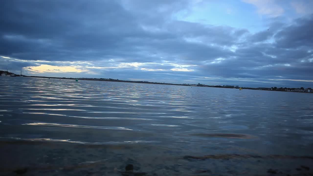 Bay waters sway peacefully; small waves reflect raising or setting sun; water looks cool and calm. Sky is blue. Camera is at a low angle. Footage in HD