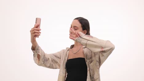 A young woman takes selfies with her smartphone, showing various hand gestures like a peace sign, blowing a kiss, and thumbs up, against a white background