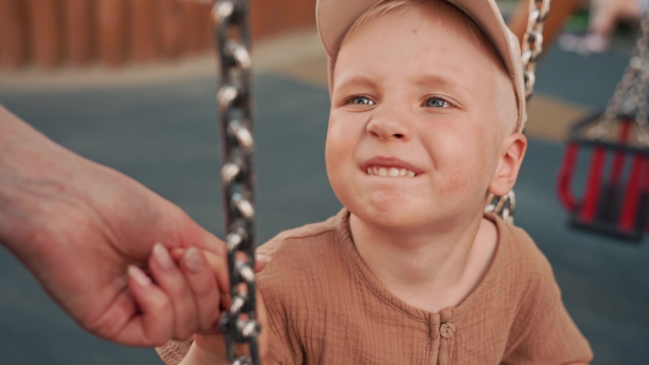 Un bambino è seduto sull'altalena in un parco giochi, spinto delicatamente da una mano adulta. La scena cattura una tenera interazione e una calma osservazione del gioco all'aperto, con una messa a fuoco morbida su mani e altalene.