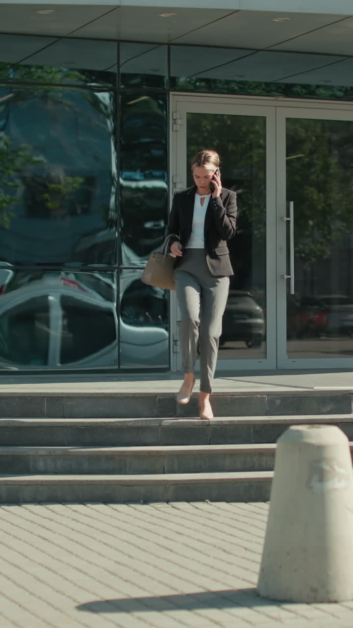 Young businesswoman in formal outfit on phone call descending stairs outside modern office building with glass reflections of pedestrians and car passing by