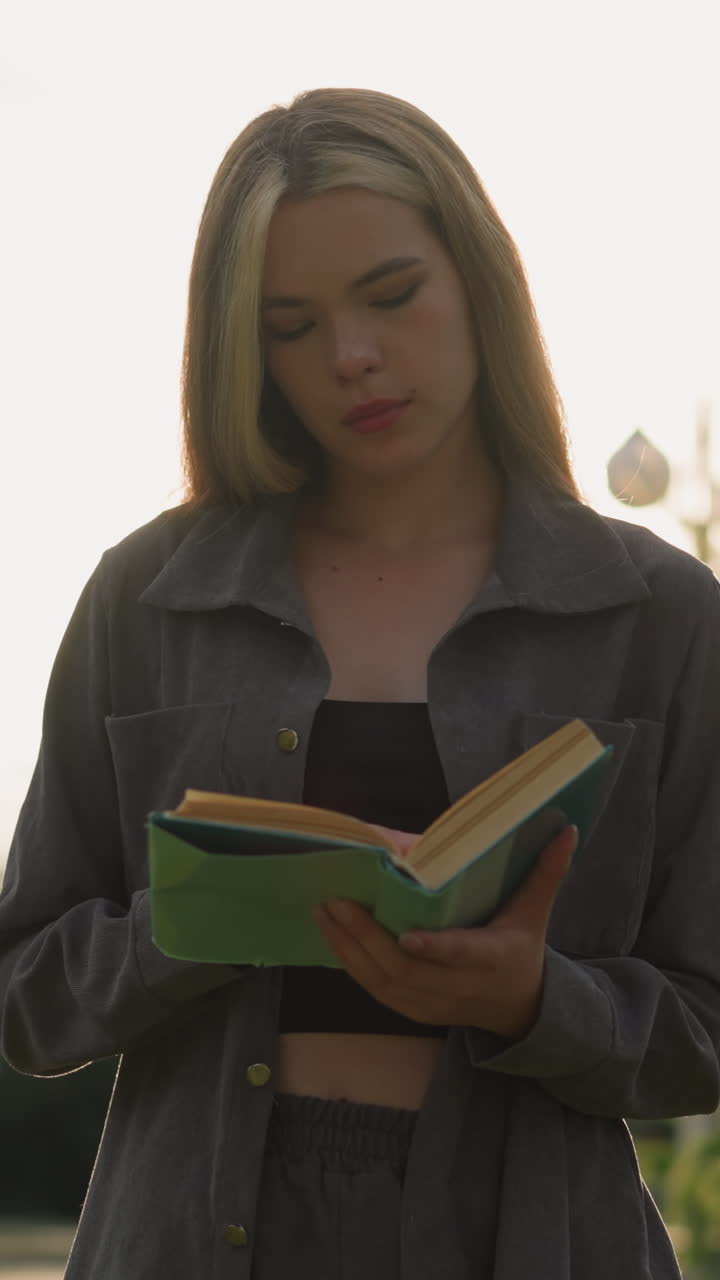 dama con ropa gris de pie al aire libre con la cabeza inclinada ligeramente volviendo las páginas de un libro, la luz del sol reflejándose suavemente a su alrededor como fondo, con árboles borrosos y edificios
