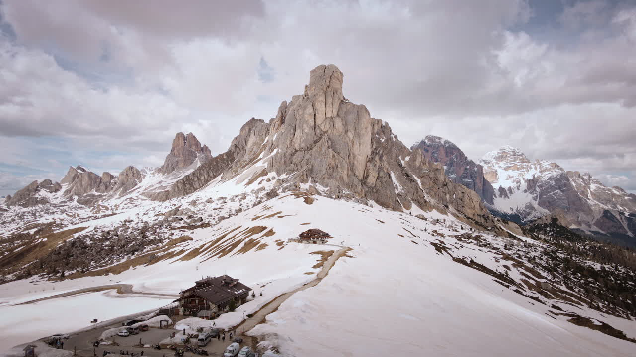 Camera glides sideways above snow-streaked slopes and a mountain hut, panning along the jagged Dolomite ridge as distant peaks and valleys slowly come into view