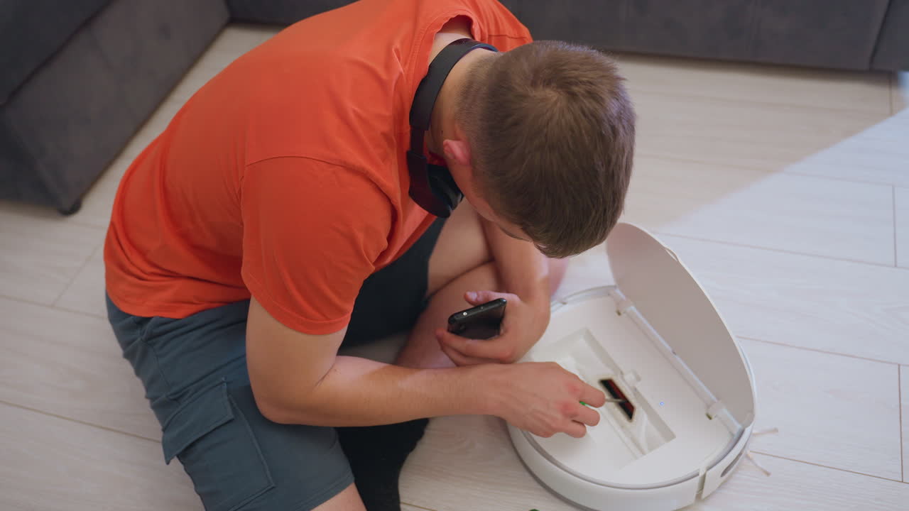 Vacuum repairer in orange shirt seated on floor beside sofa inspects robot vacuum cleaner using screw while holding phone, focused on maintenance and repair of smart cleaning device in home interior